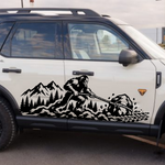 A white SUV parked outdoors on a paved lot, featuring a large black mountain-and-sasquatch silhouette decal running along the side door.