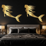 A modern bedroom featuring a black wall with two large gold mermaid wall decals above a bed dressed in black bedding. The room is styled with matching black nightstands, lit table lamps, and a potted plant.