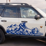 A white SUV with a blue vinyl decal of a sasquatch/bigfoot pulling a boulder with a mountain scene in the background. T