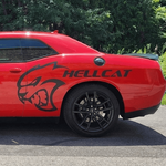 A red Dodge Challenger Hellcat sports car parked outdoors on a paved road with green trees and blue sky in the background. The side of the car features a black Hellcat cat head graphic and the word 'HELLCAT' in black.