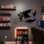 A styled home office or gaming room with a dark gray accent wall featuring a large black Sonic the Hedgehog silhouette decal. The room includes shelves filled with video game cases, a small Sonic-themed bookshelf, a brown leather office chair, a computer desk setup, and framed wall art.