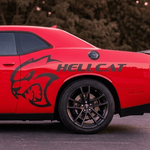 A red Dodge Challenger with black wheels and a large black Hellcat graphic decal on the side, parked outdoors on asphalt with trees in the background.